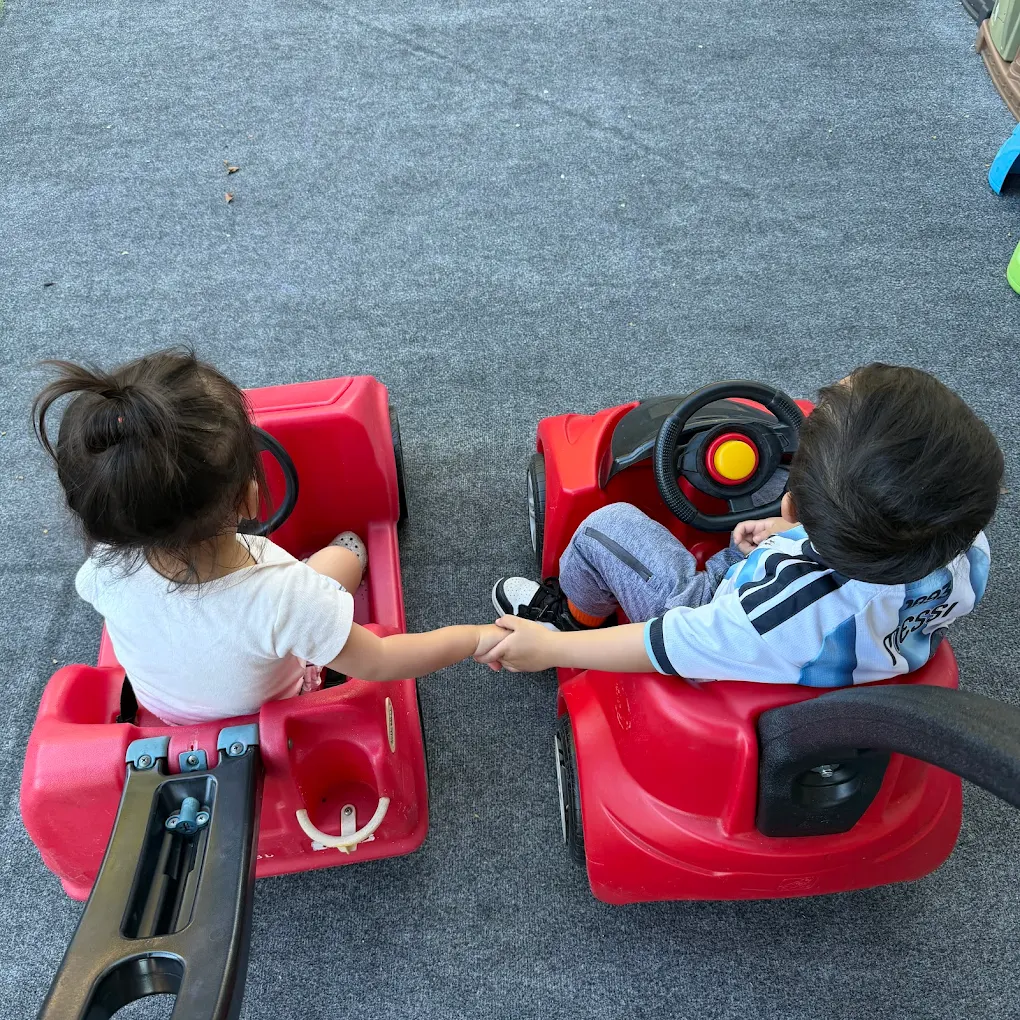 Two toddlers holding hands while riding red ride-on cars at Crayoland