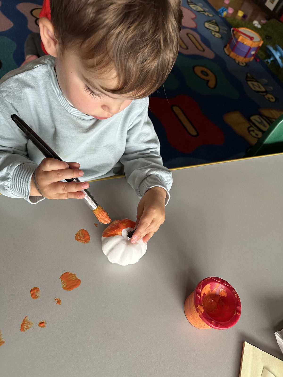 Toddler painting a small white pumpkin orange during a fall art activity